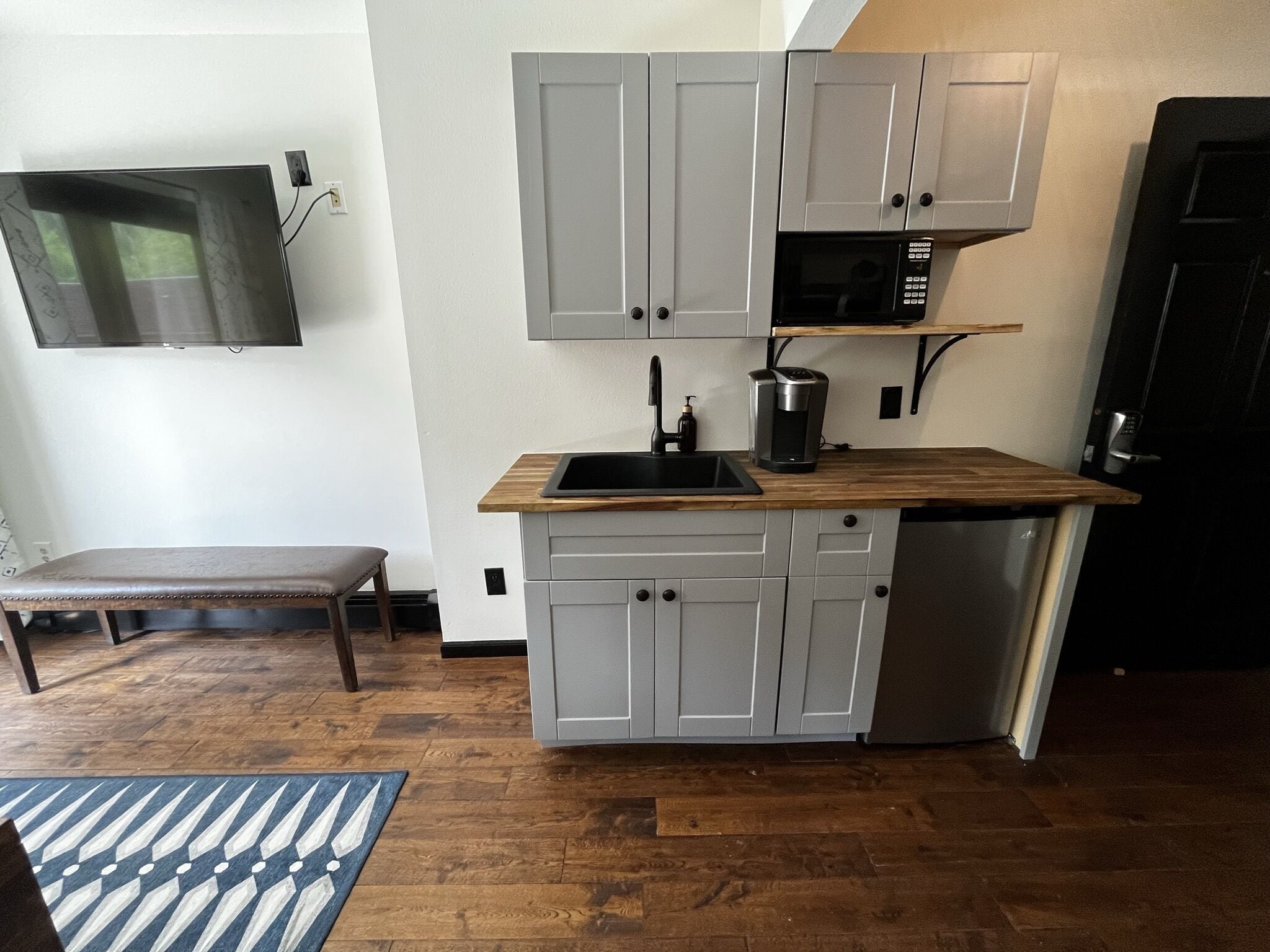 A compact kitchen with grey cabinets, a wooden countertop, a black microwave, and a built-in dishwasher.