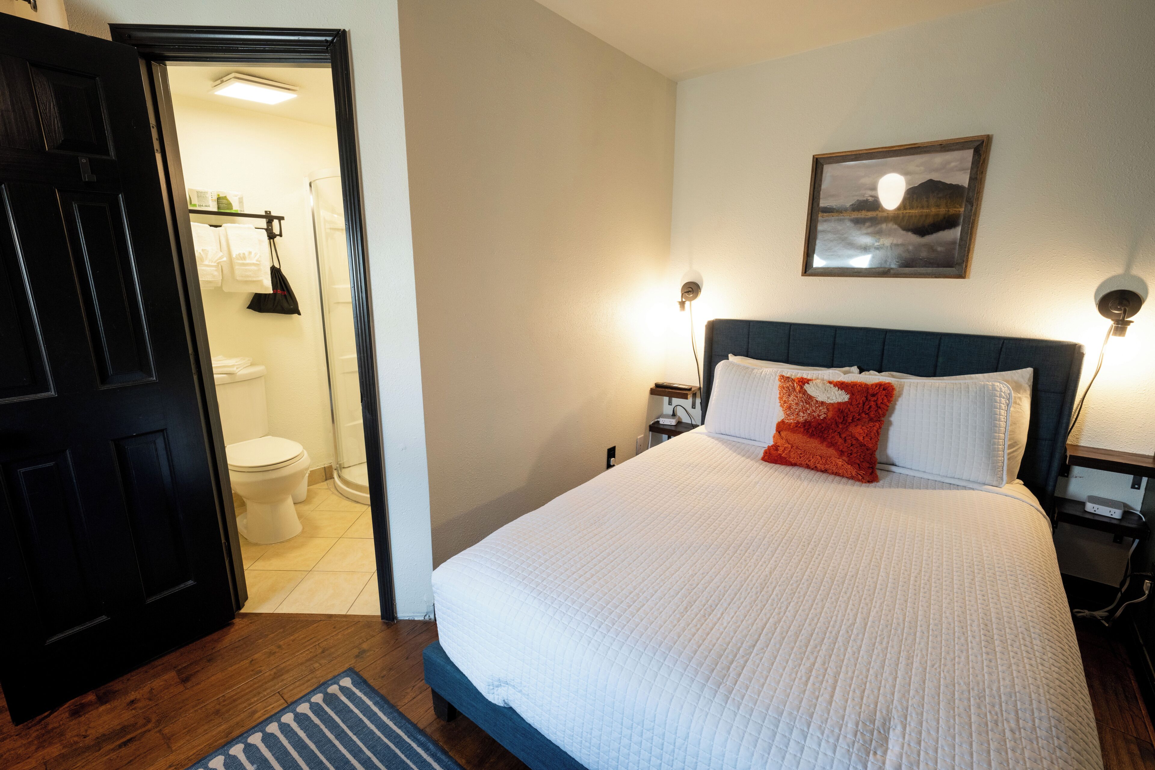 Habitación de hotel con cama, mesita de noche, lámpara de pared, un cuadro y un baño visible a través de una puerta abierta.