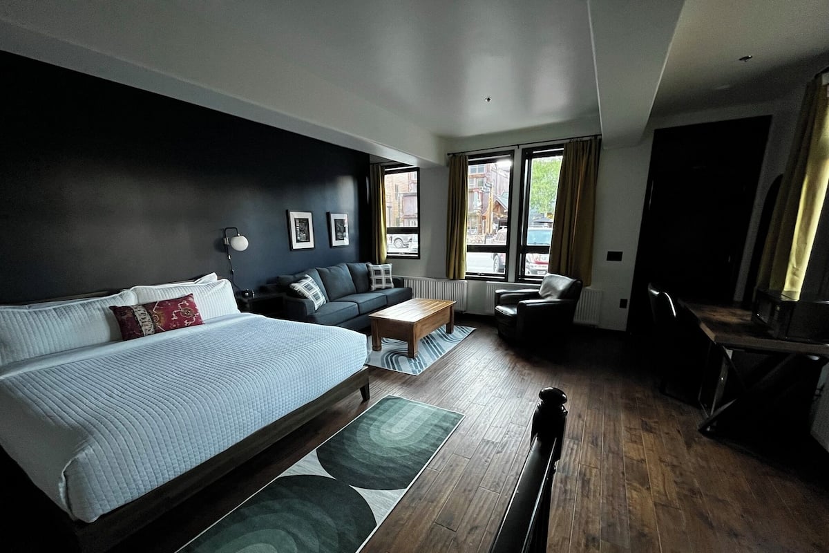 A modern hotel room with a large bed, a sofa, a dining table, and a chair. There are windows with curtains, a radiator, and a view of the street outside.
