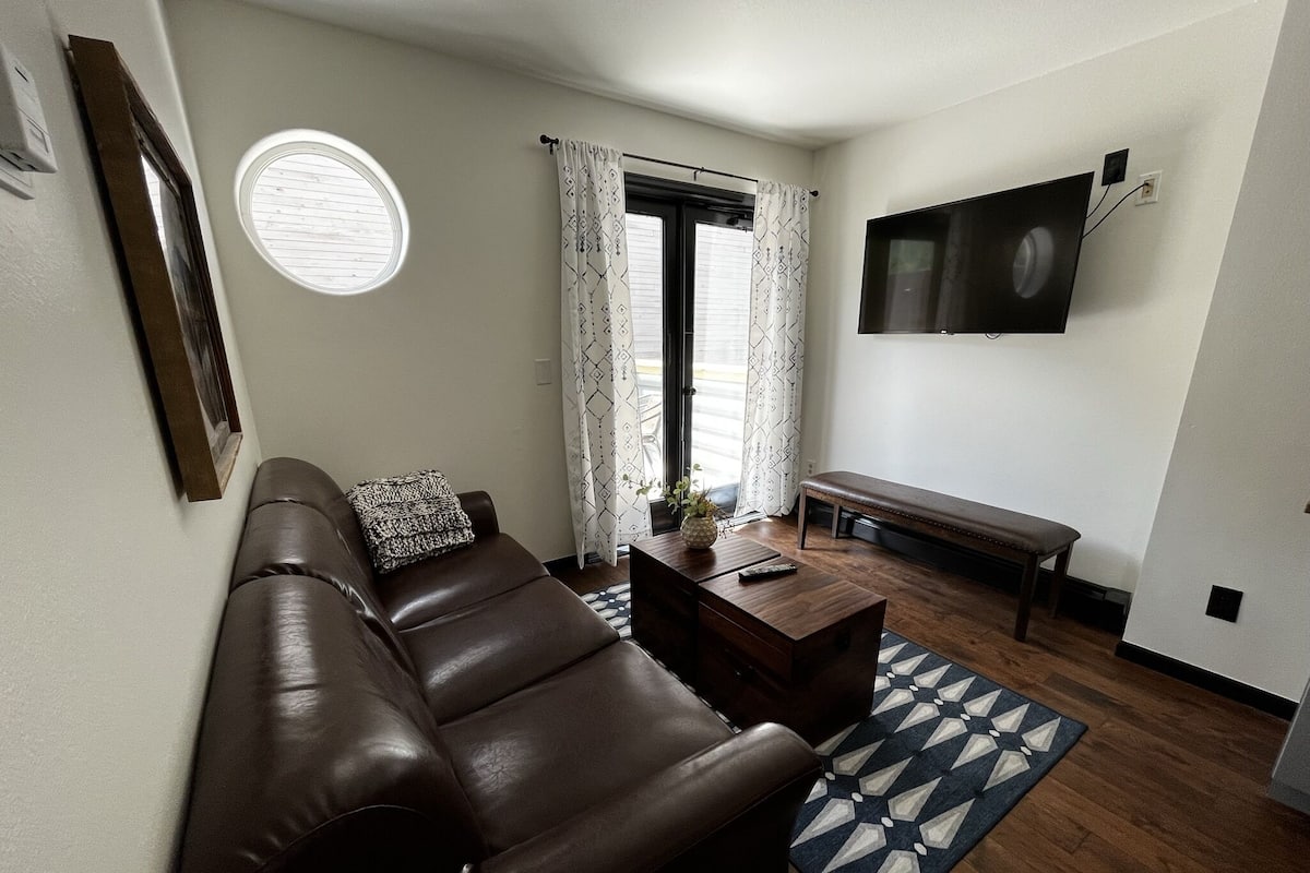 A living room with a brown leather sofa, a wooden coffee table, and a bench. There is a TV mounted on the wall, a window with curtains, and a patterned rug on the floor.