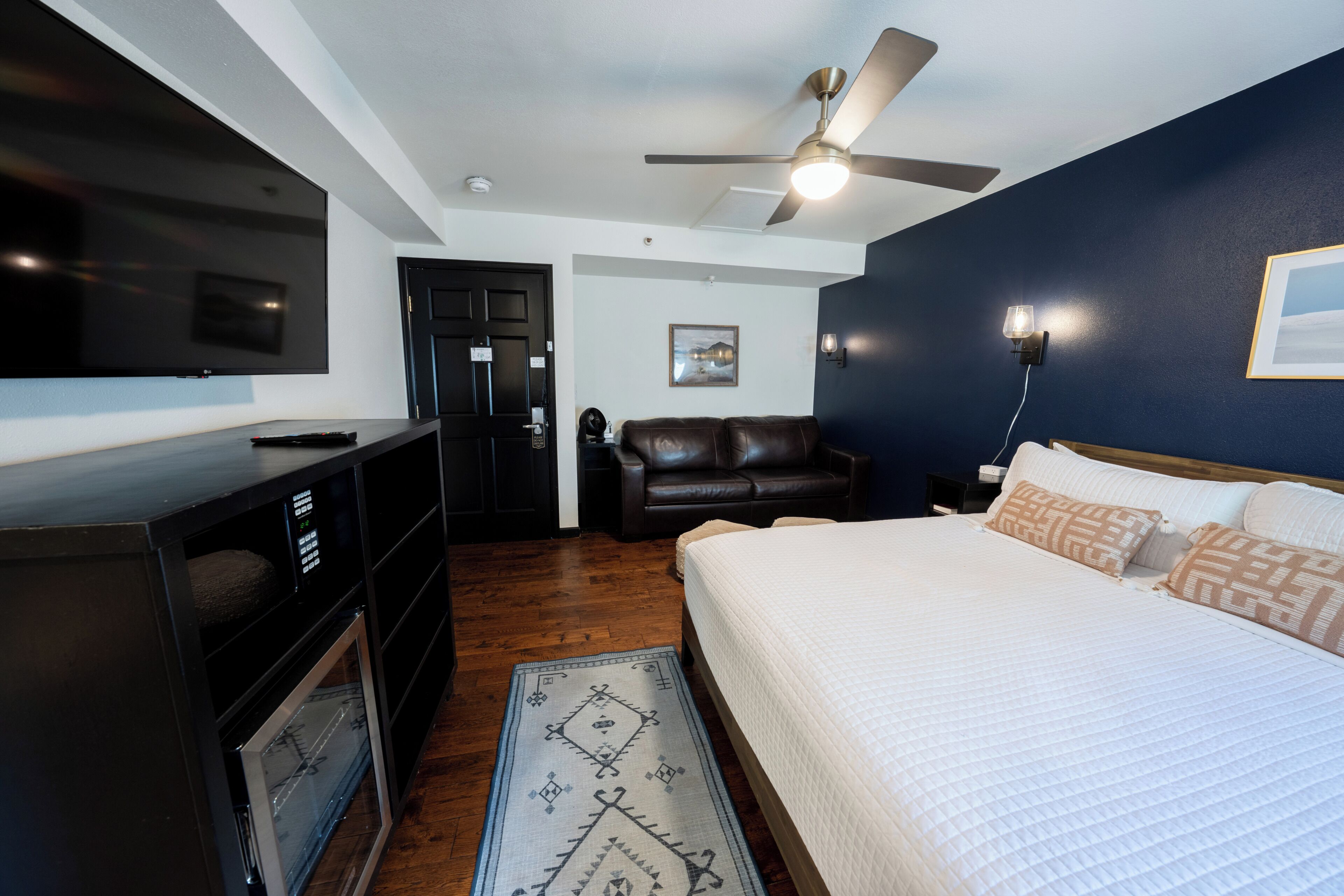 A hotel room with a bed, a TV, a ceiling fan, a desk, and a door.