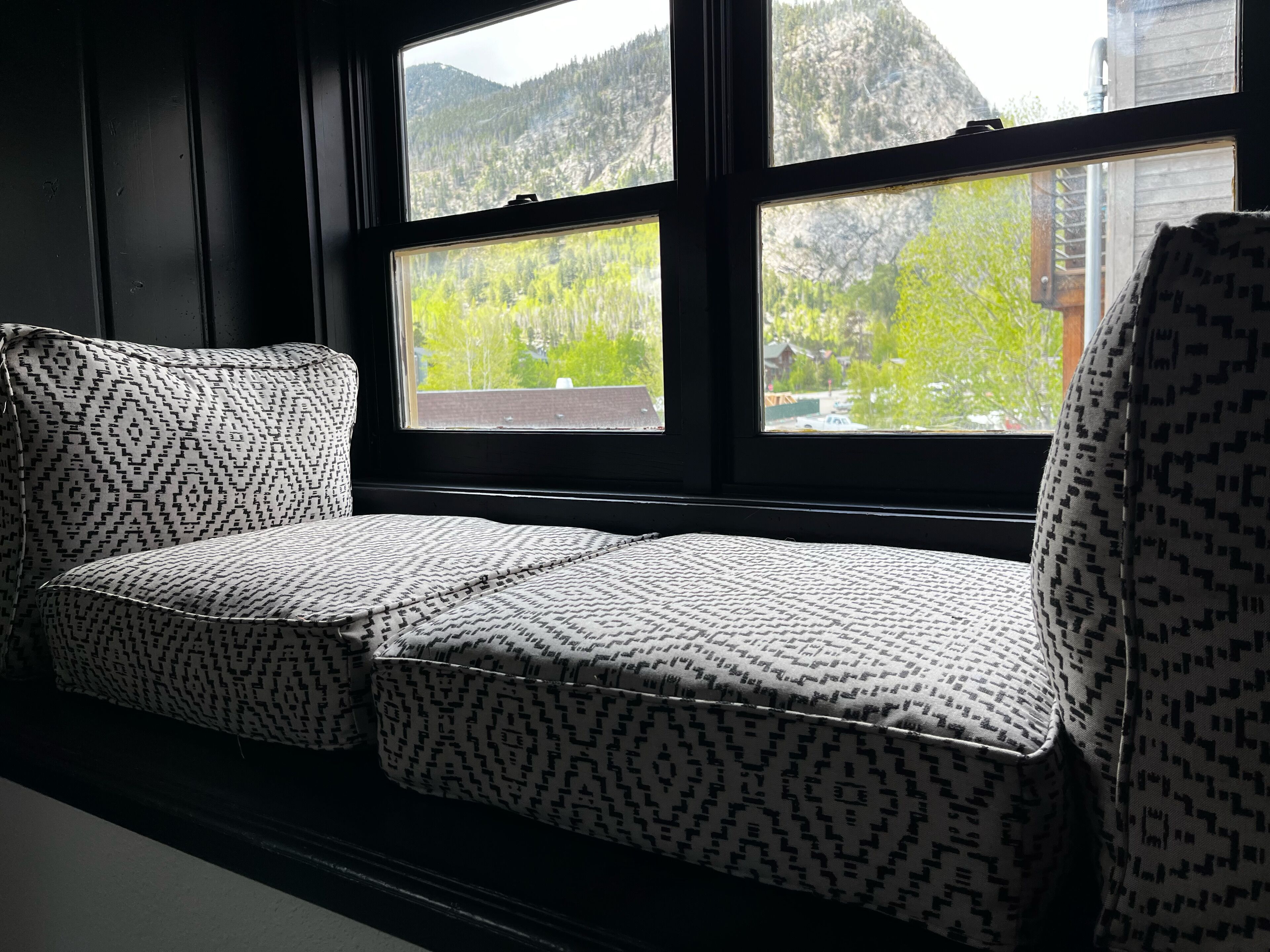 Un asiento junto a la ventana con cojines estampados, vistas a una montaña boscosa y un edificio al fondo.