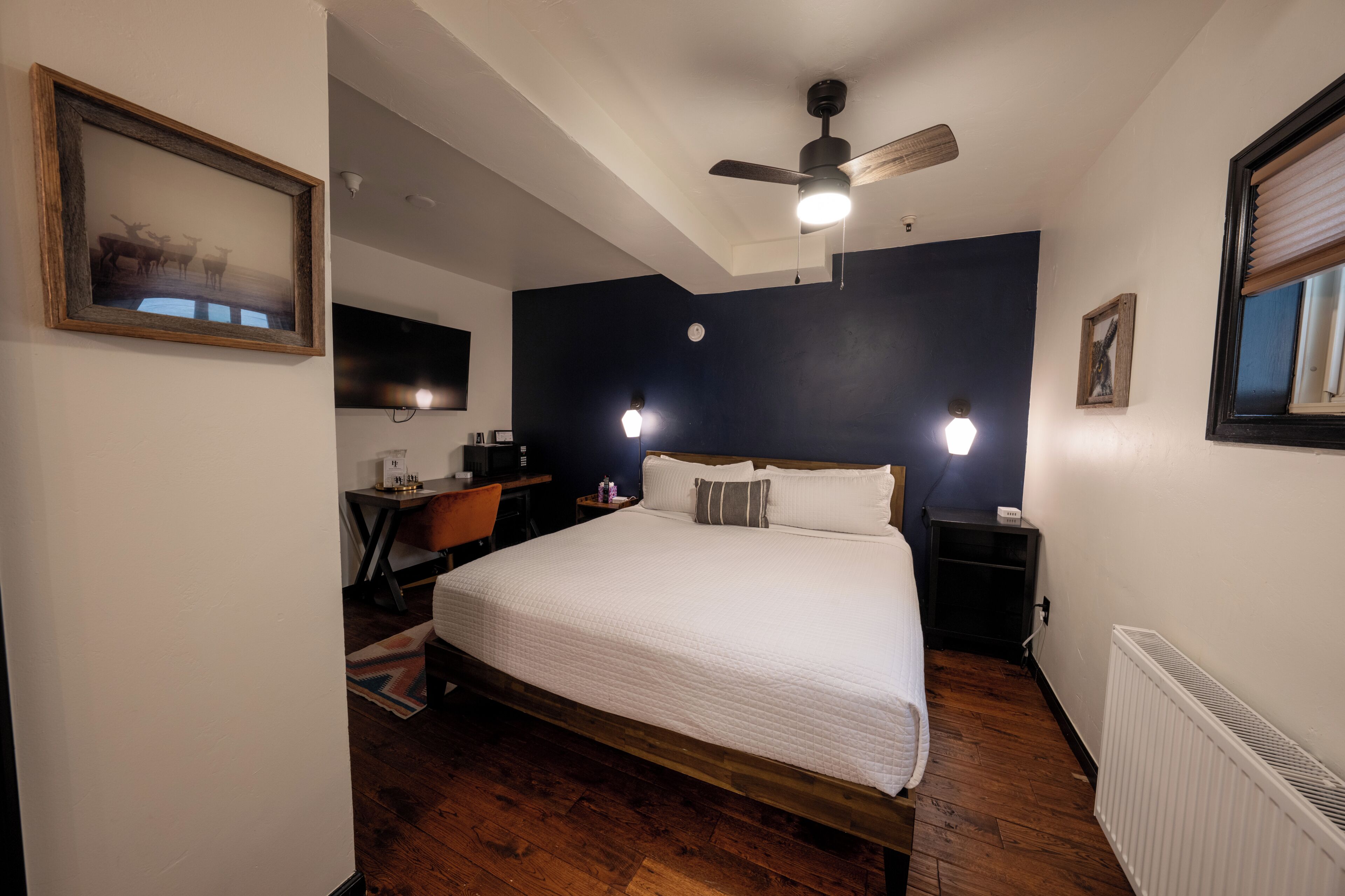 A bedroom with a bed, a desk, a chair, a ceiling fan, and a wall-mounted TV.
