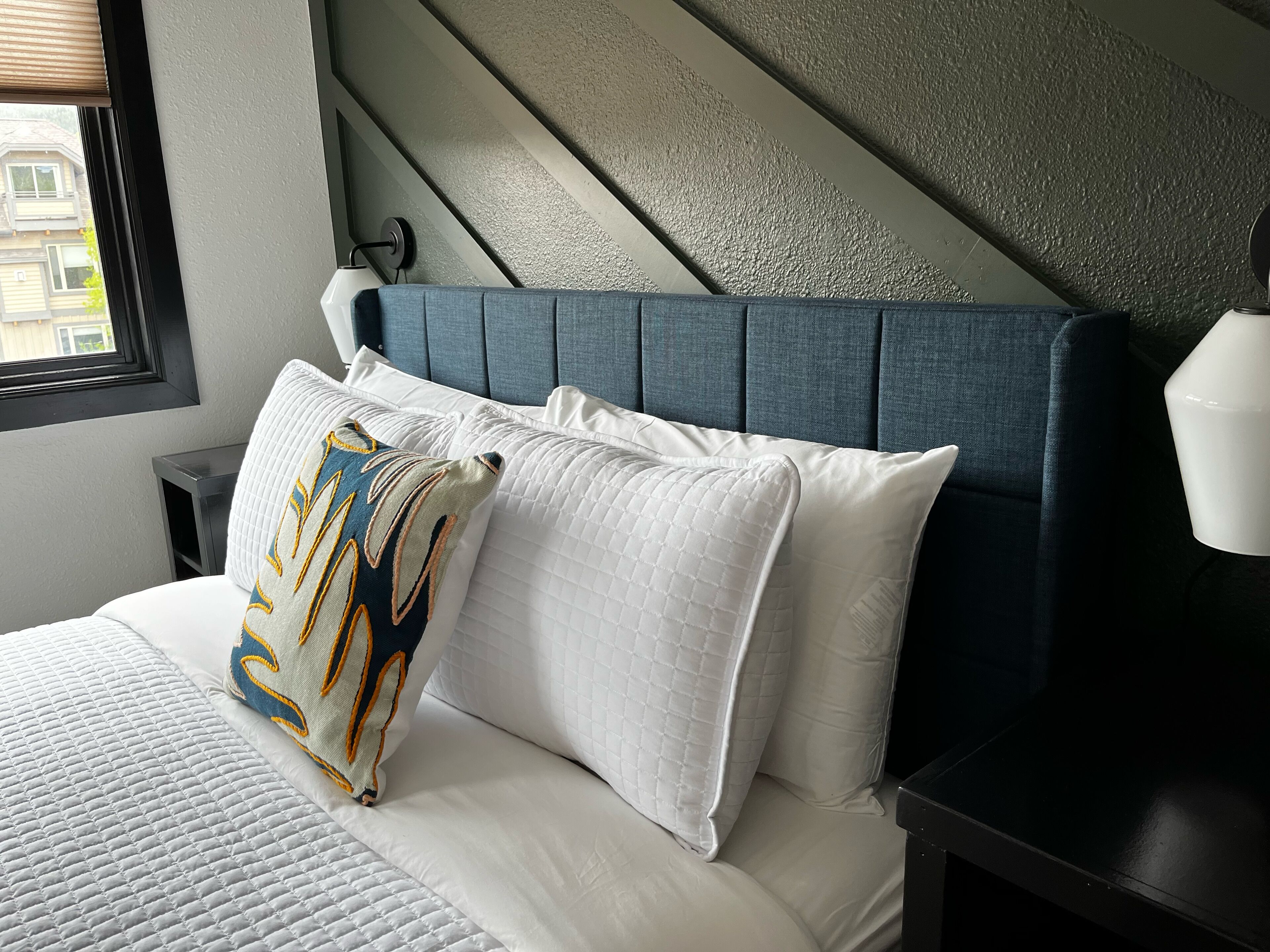 Una cama bien hecha con ropa blanca y una almohada estampada, un cabecero azul oscuro y una mesita de noche con una lámpara.