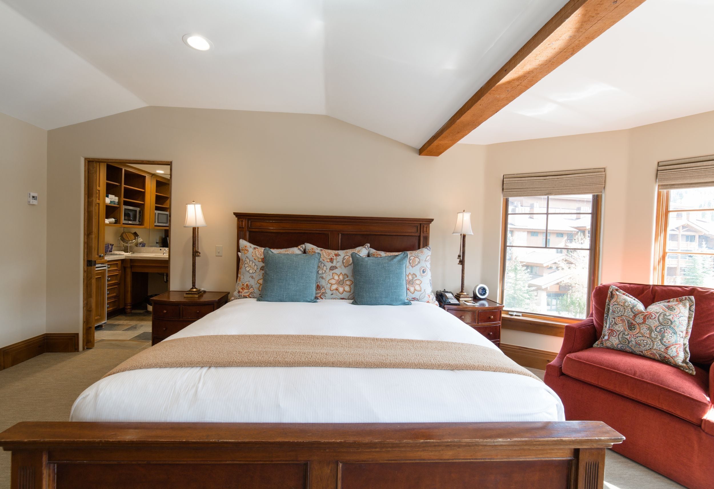 Une chambre avec un grand lit, une tête de lit en bois, des tables de chevet avec des lampes, un fauteuil rouge et une vue sur l’extérieur par une fenêtre.