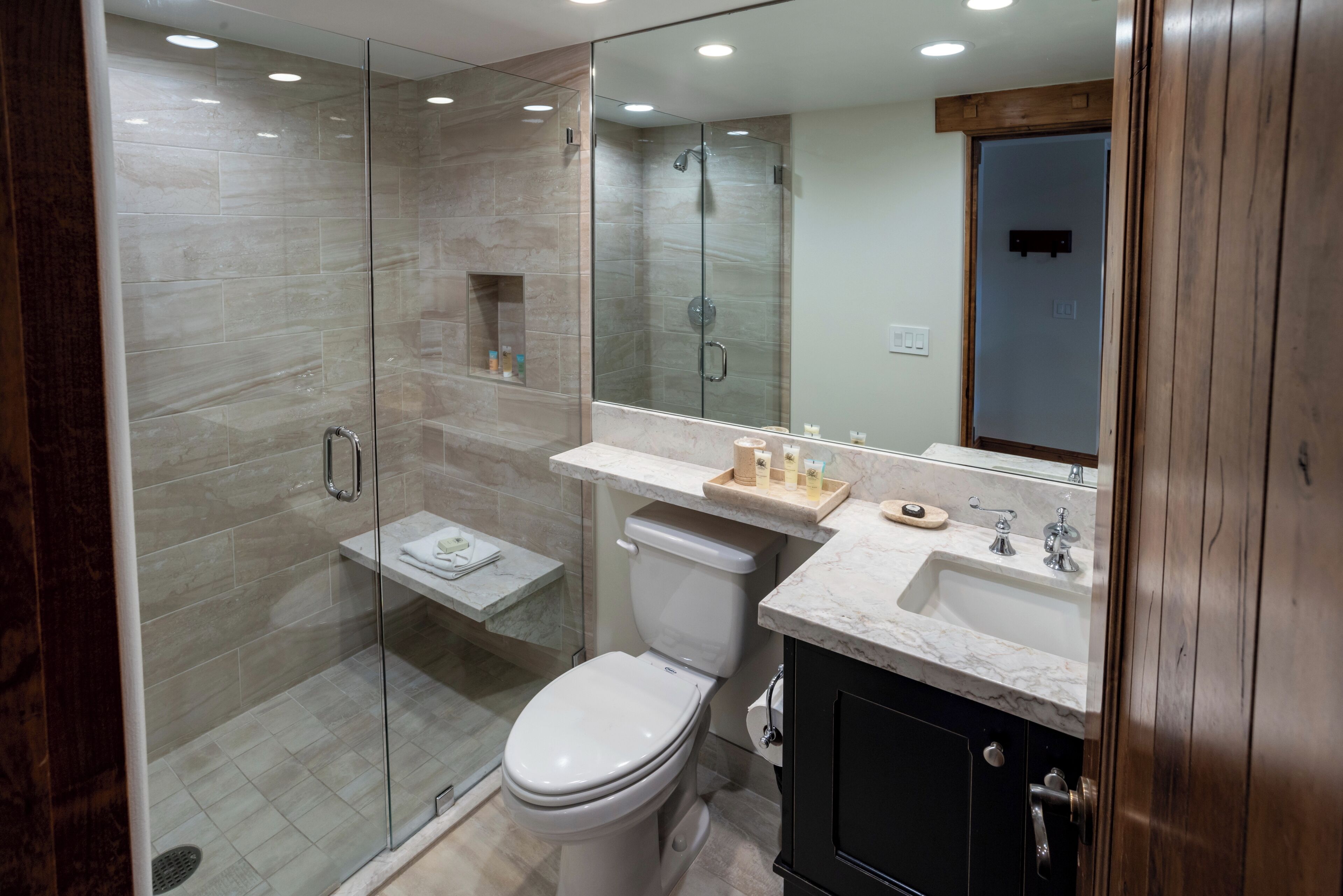 Un baño moderno con ducha envuelta en vidrio, inodoro blanco, tocador con lavamanos y espejo, y un mueble de madera.