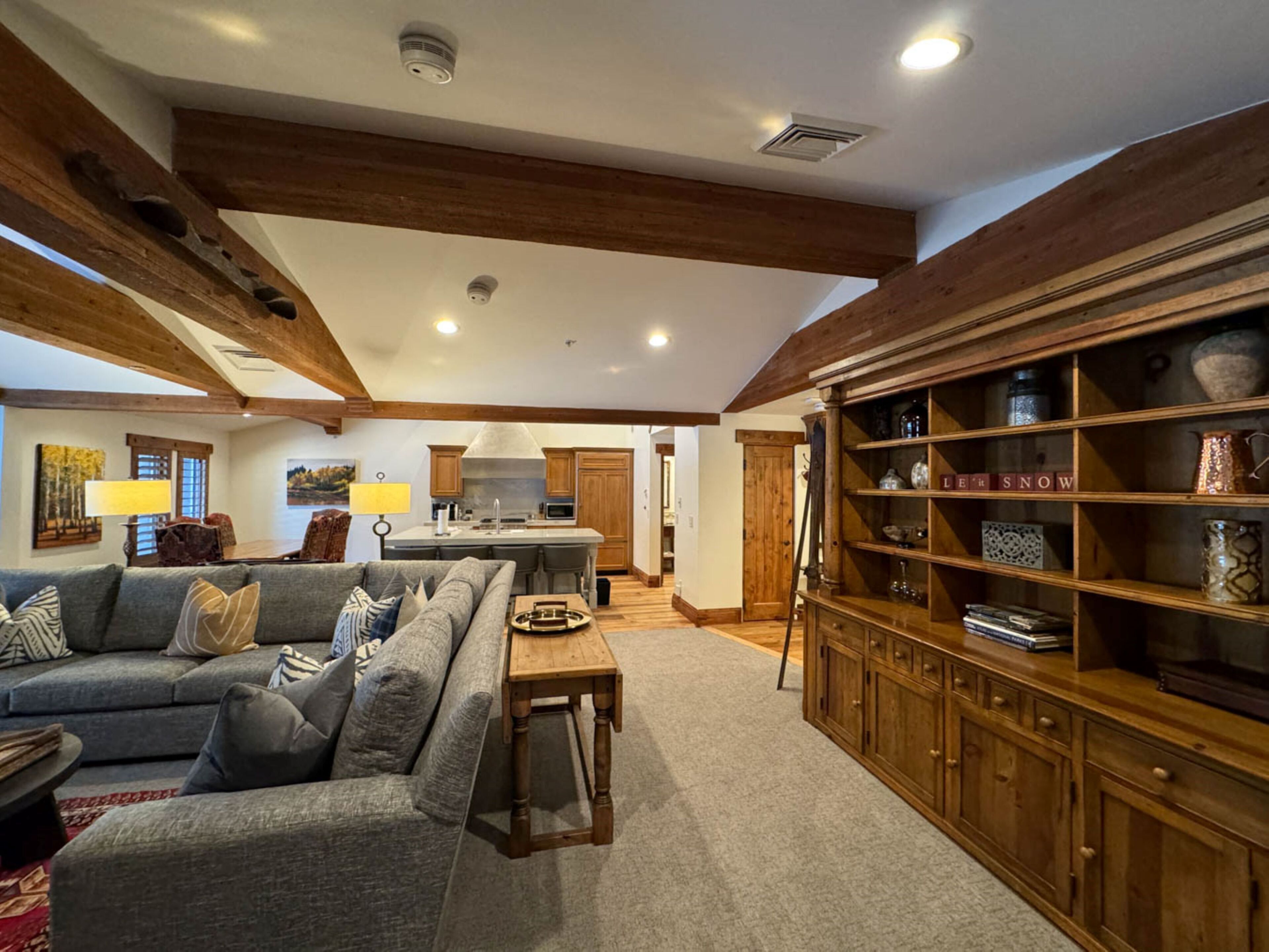 A spacious living room with a large gray sectional sofa, wooden beams on the ceiling, a kitchen area with a central island, and a wooden cabinet with decorative items.