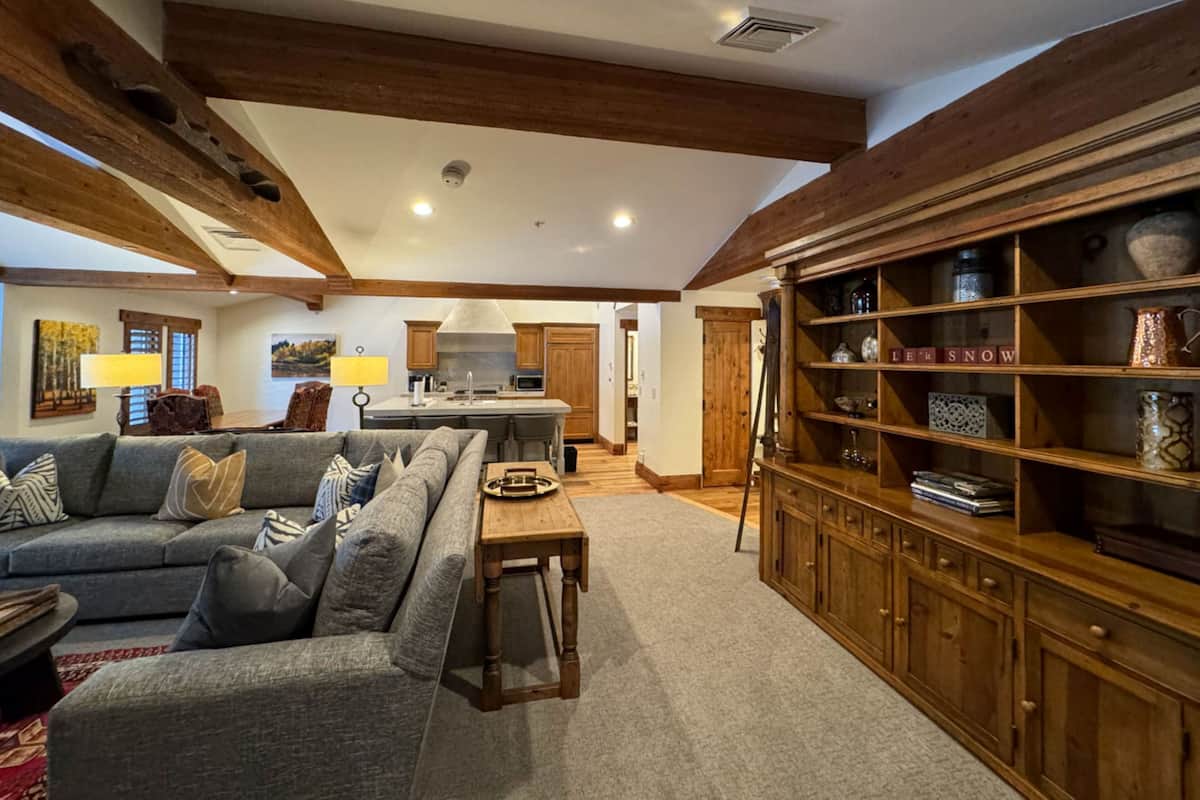 Ein geräumiges Wohnzimmer mit einer großen, grauen Eckcouch, Holzbalken an der Decke, einer Küchenzeile mit Kochinsel und einem Holzschrank mit dekorativen Accessoires.