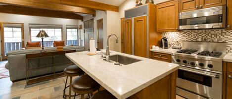 Una cocina con una isla central, electrodomésticos de acero inoxidable, armarios de madera y una zona de estar con sofá y sillas.
