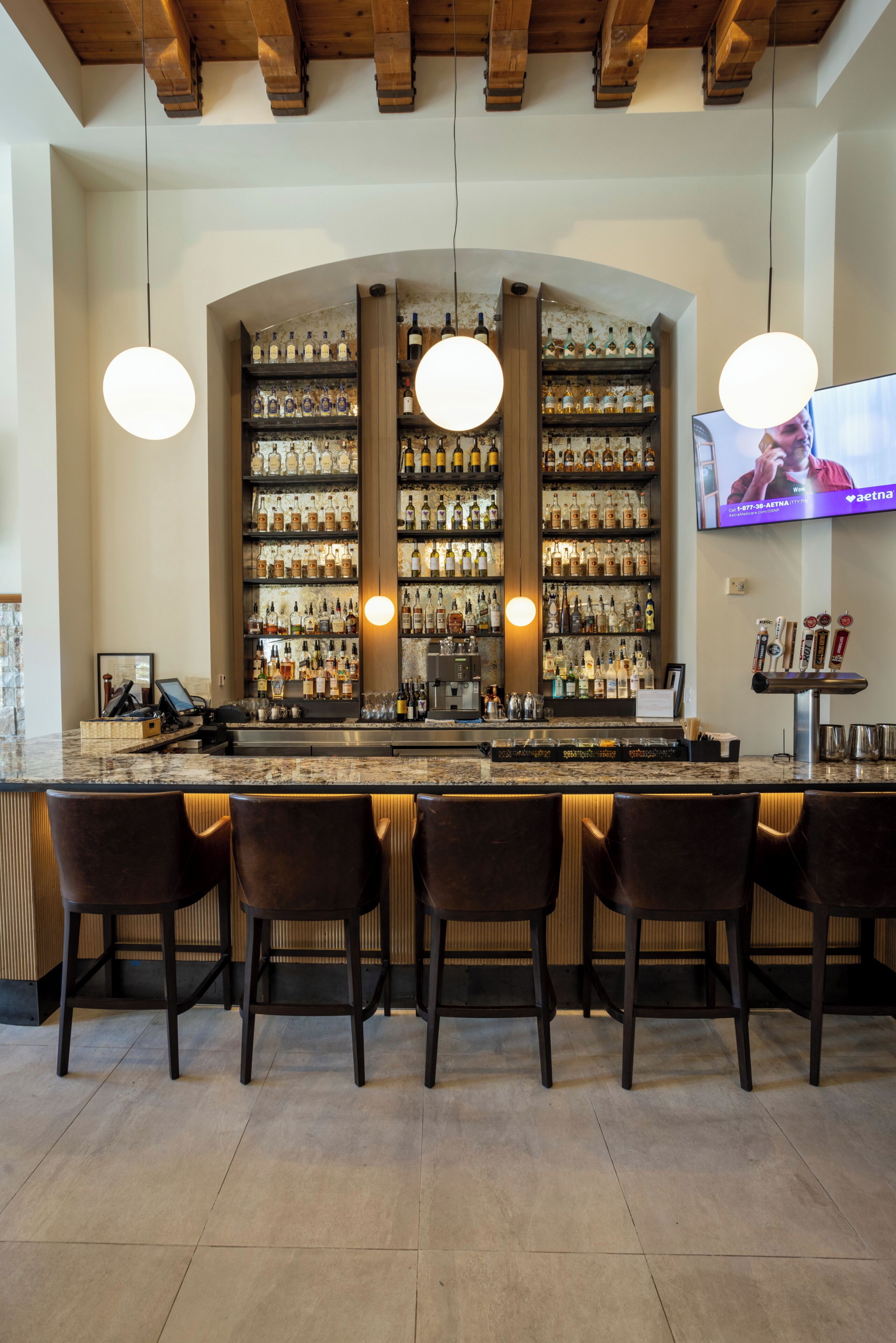 Uma área de bar com bancada de mármore, várias prateleiras cheias de garrafas, luminárias pendentes e uma tela de televisão.