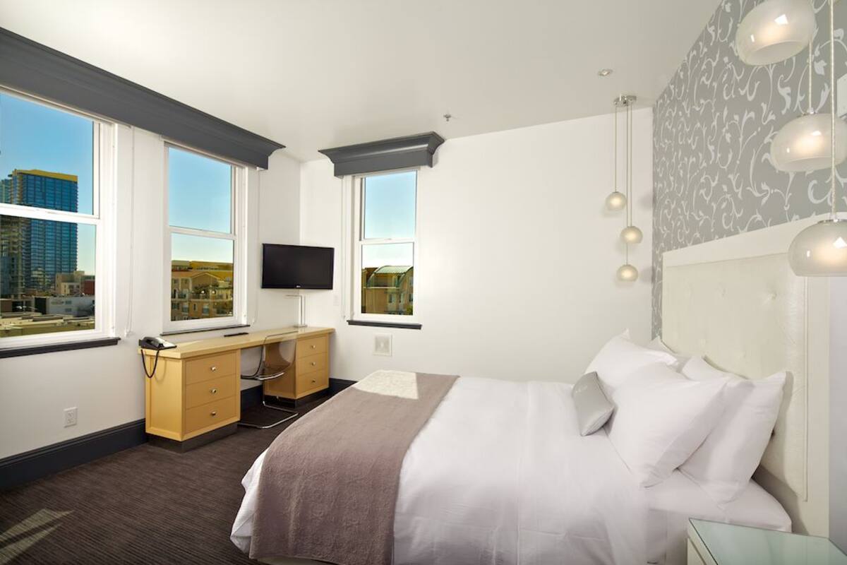 Quarto de hotel com cama, escrivaninha e duas janelas com vista para a cidade.