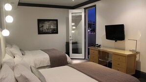 A hotel room with two beds, a desk, a TV, and a city view through a glass door. - La Pensione Hotel (San Diego)