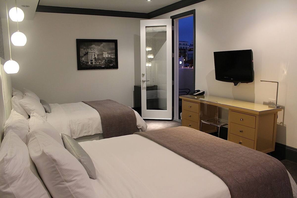 Quarto de hotel com duas camas, uma mesa, uma televisão e vista para a cidade através de uma porta de vidro.