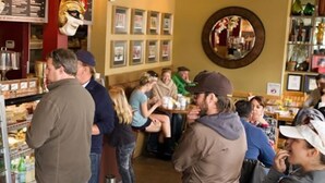 A busy cafe with people standing and sitting at tables, a counter with various food items, and shelves with products. - La Pensione Hotel (San Diego)