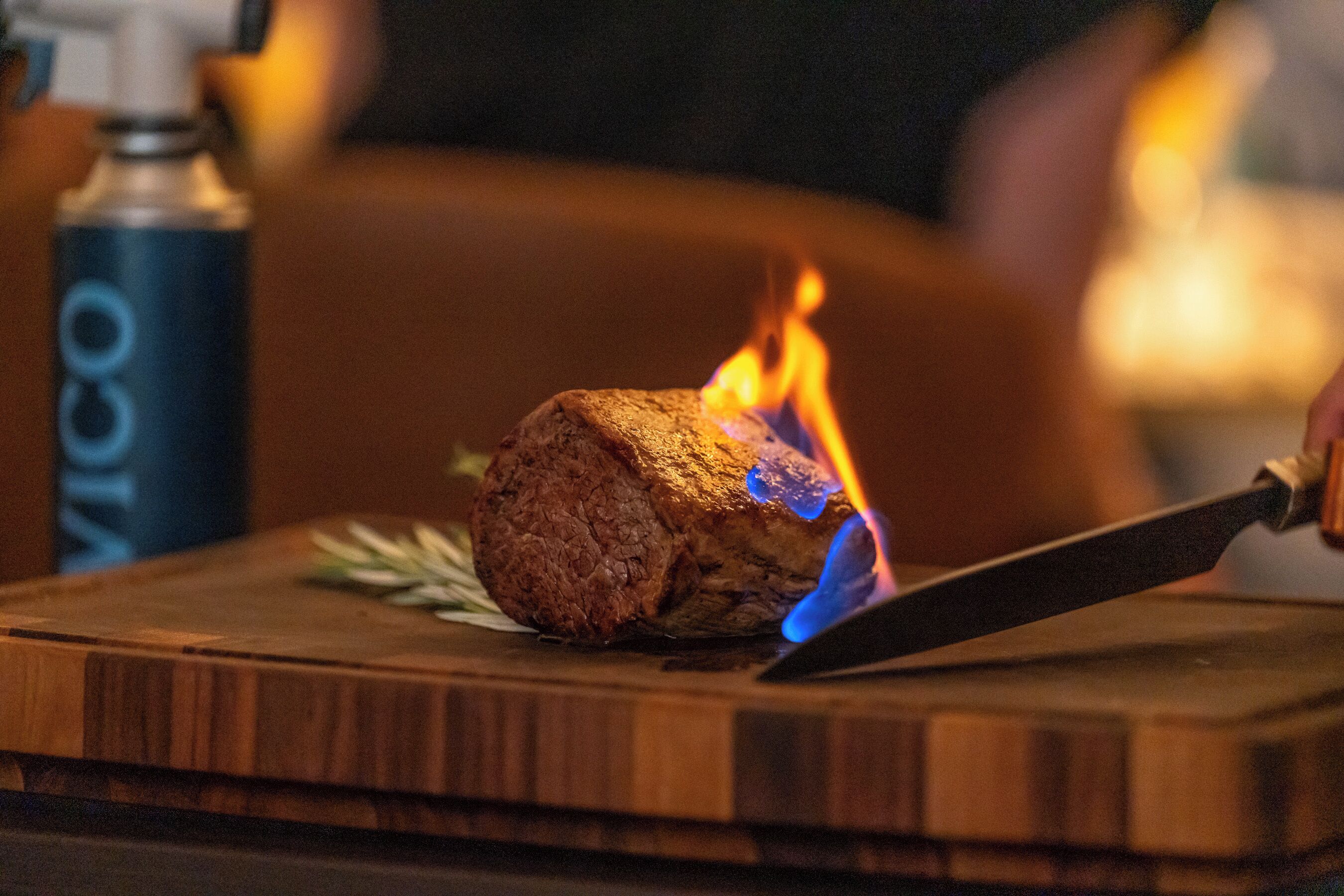 Una tabla de madera con un asado en una parrilla, un cuchillo y una botella de VICO al fondo.