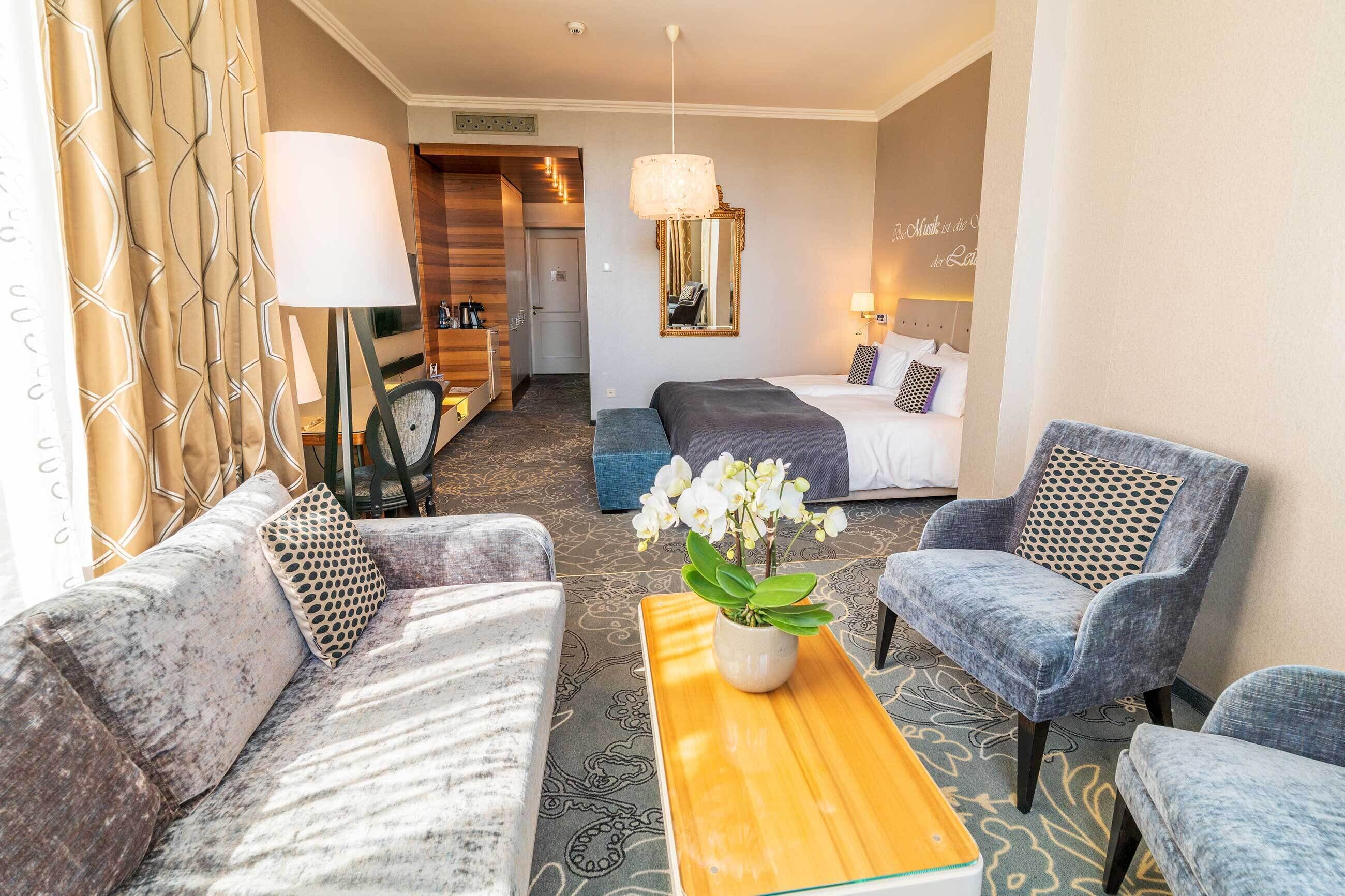 Een hotelkamer met een bank, fauteuil, een houten bijzettafeltje met een plant, een bed met hoofdeinde, een spiegel en een lamp.