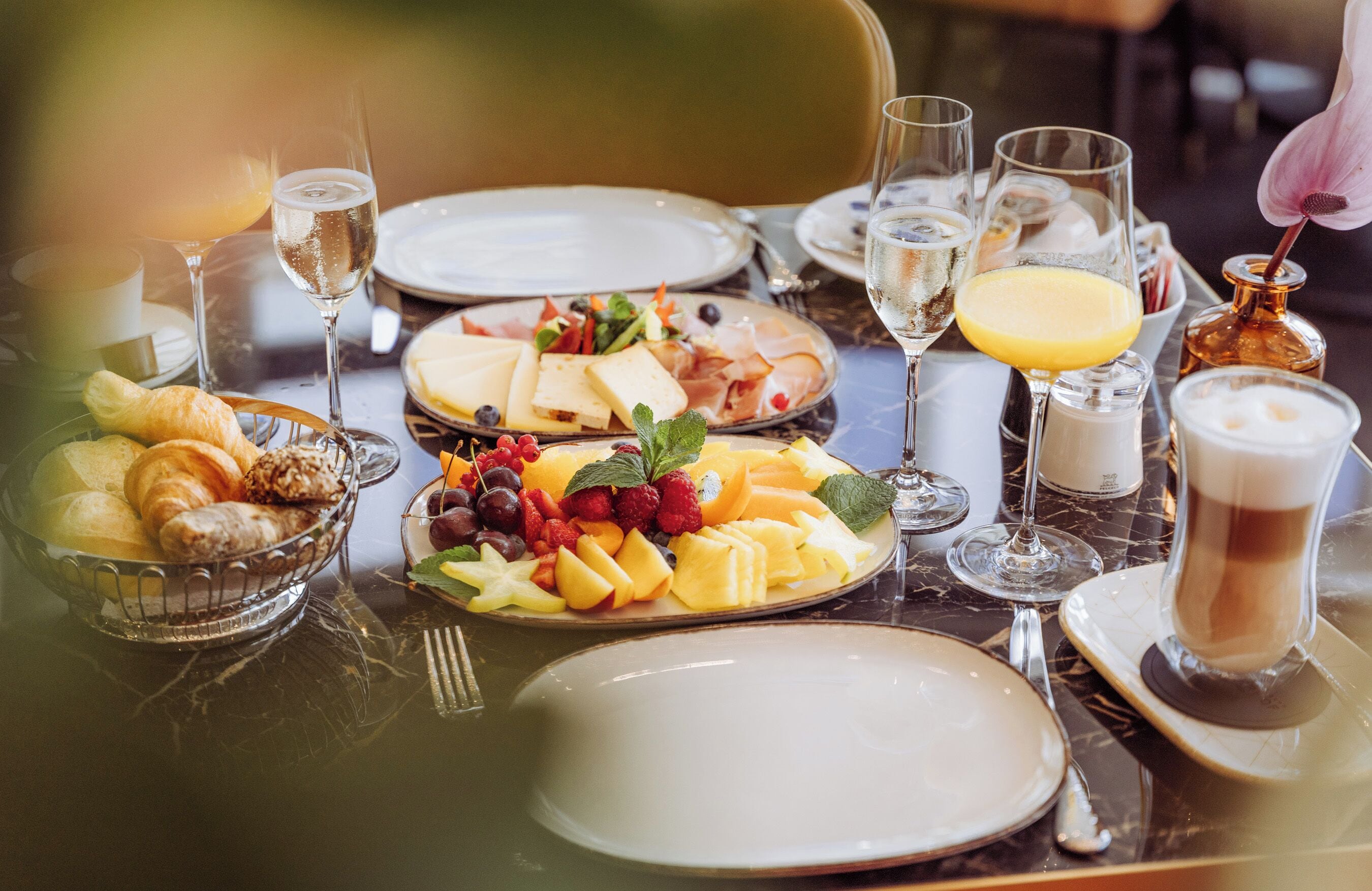 A breakfast setting with plates of fruit, pastries, and cheese, along with glasses of juice and wine.