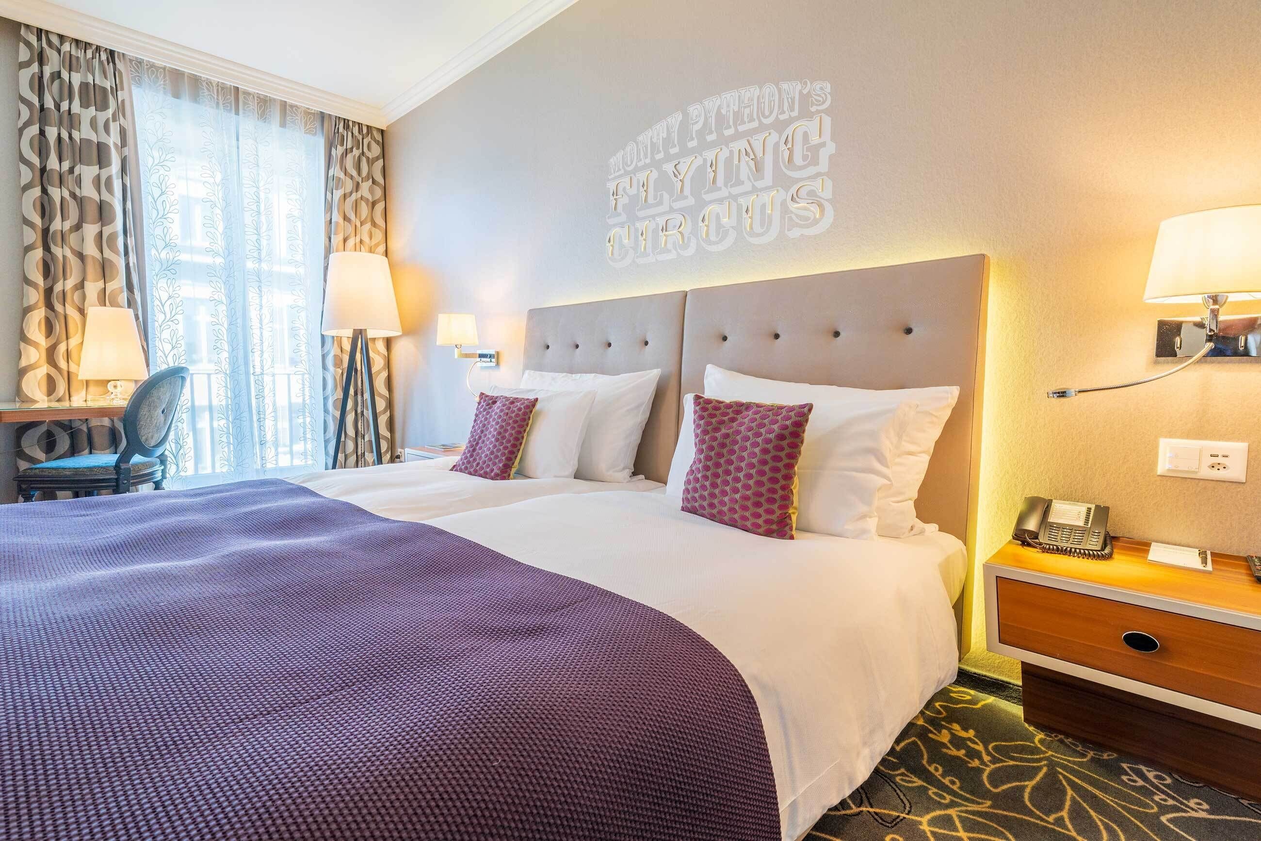 A hotel room with two beds, a desk, a chair, a lamp, a phone, and a wall sign.