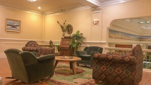 A spacious lobby with a wooden floor, patterned armchairs, a central table with a vase and decorative items, a painting on the wall, and a balcony with a view.