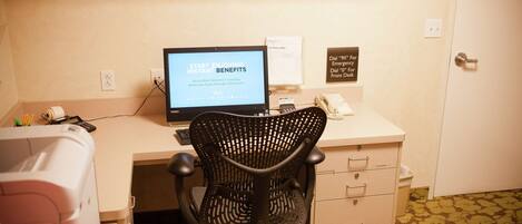 A hotel room with a desk, a computer, a chair, a printer, and a trash bin. There are signs on the wall indicating emergency and front desk phone numbers.