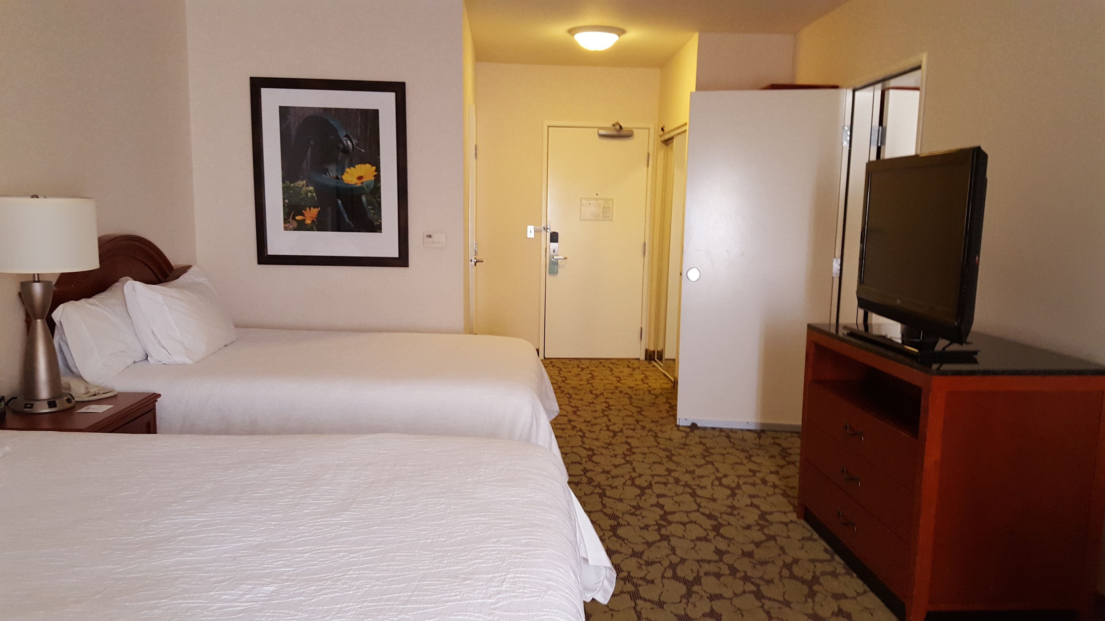A hotel room with two beds, a television on a wooden cabinet, a framed picture on the wall, and a door leading to another room.