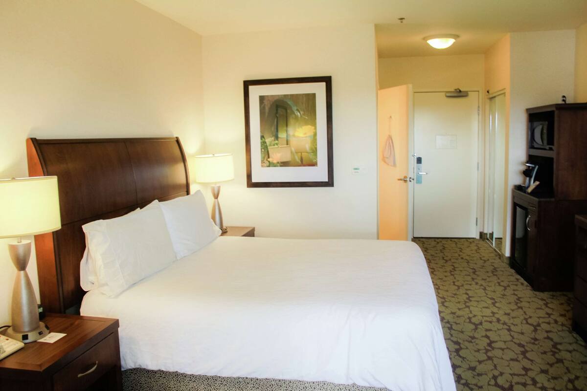 A hotel room with a large bed, two bedside lamps, a framed picture on the wall, a wardrobe, and a door leading to another room.