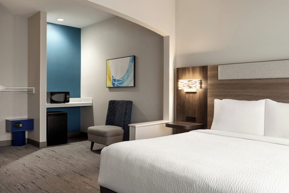 A hotel room with a bed, a chair, a desk with a microwave, a trash bin, and a wall-mounted artwork.