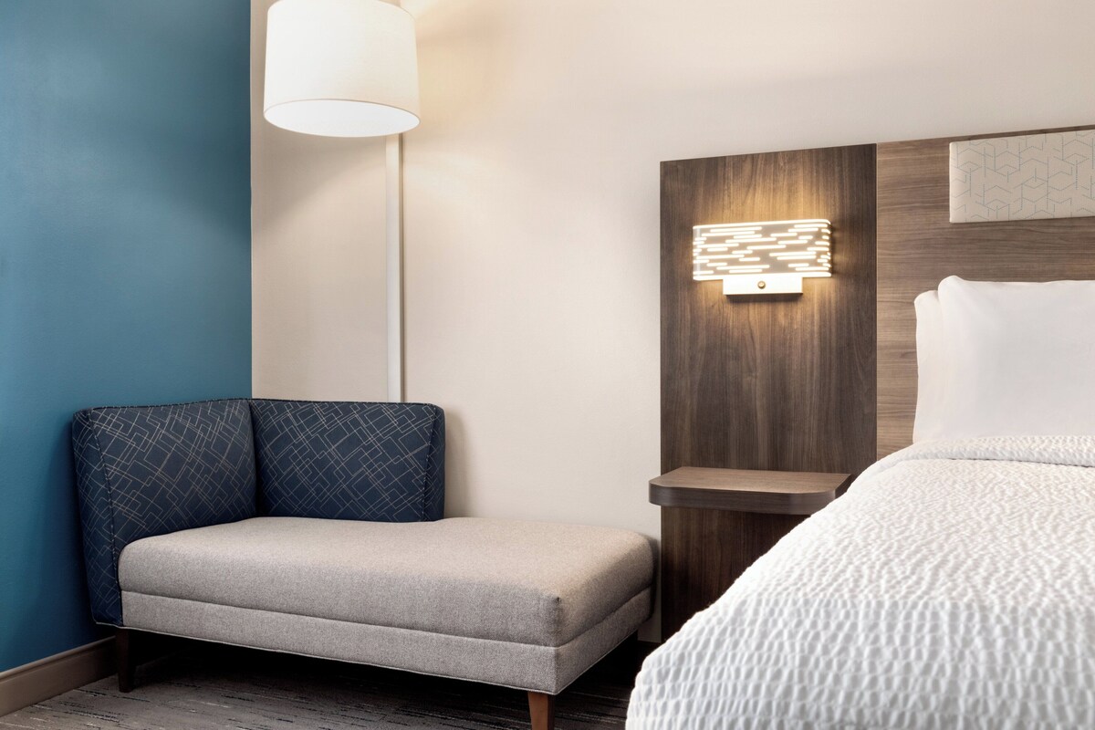 A hotel room with a sofa, a bed, a nightstand, a lamp, and a wall-mounted light fixture.