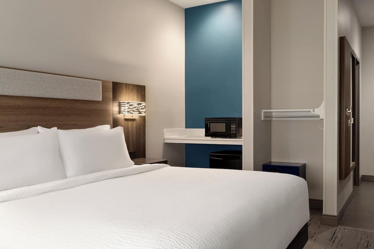 A modern hotel room with a large bed, a bedside table with a lamp, a microwave, and a small blue cabinet.