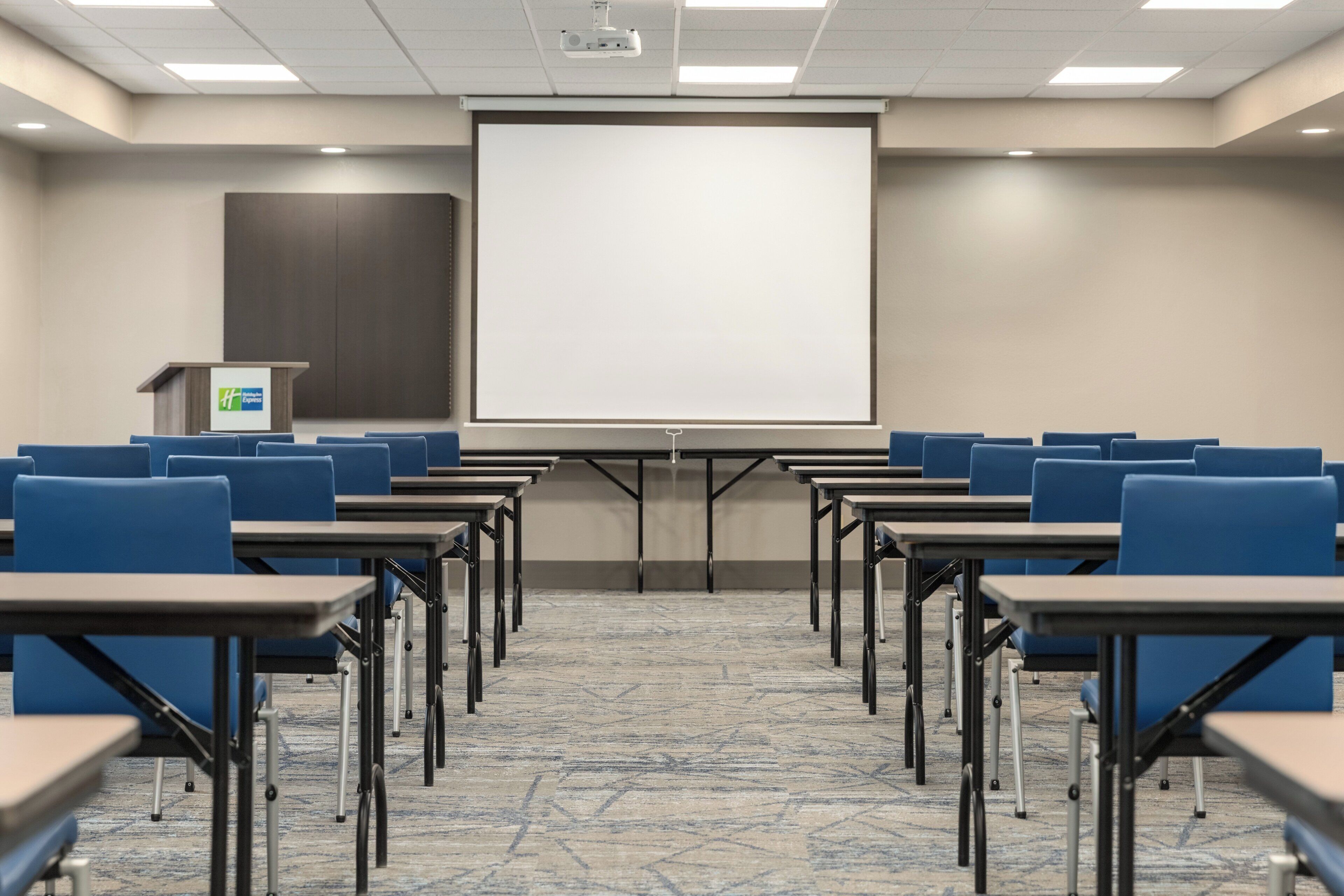 Un salón de conferencias con filas de sillas azules, un atril y una gran pantalla de proyección.