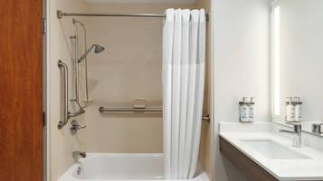 Une salle de bain avec une baignoire, une douche équipée de barres d’appui, un rideau blanc, un lavabo avec un miroir et des articles de toilette posés sur le comptoir.