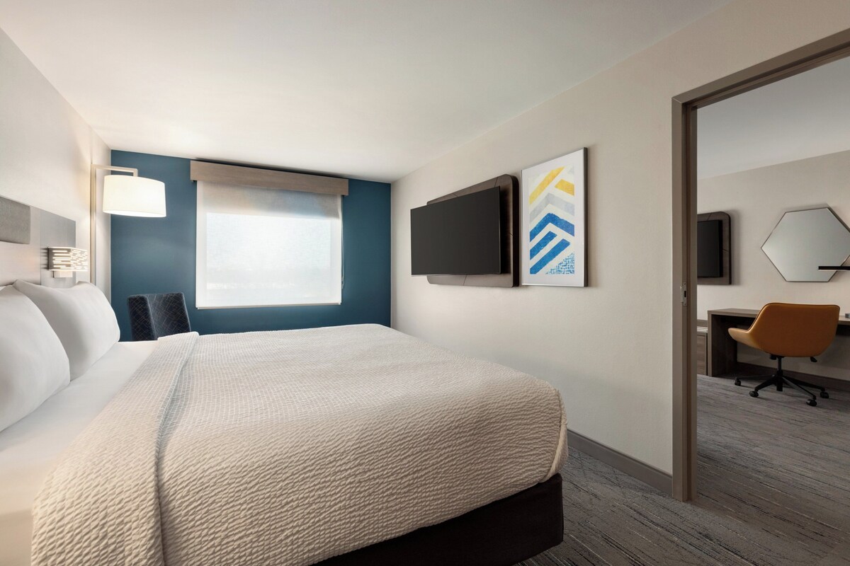 A hotel room with a bed, a desk, a chair, a TV, and artwork on the wall.