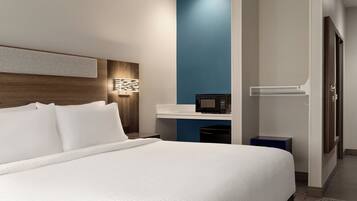Una habitación de hotel moderna con una cama grande, una mesita de noche con lámpara, un microondas y un pequeño armario azul.