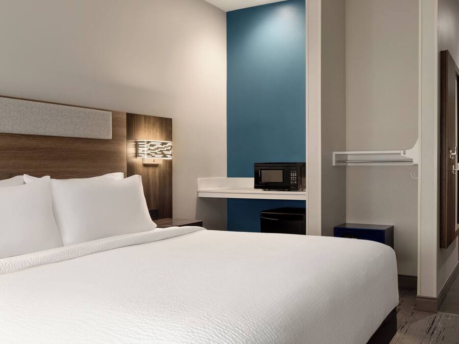 Una habitación de hotel moderna con una cama grande, una mesita de noche con lámpara, un microondas y un pequeño armario azul.