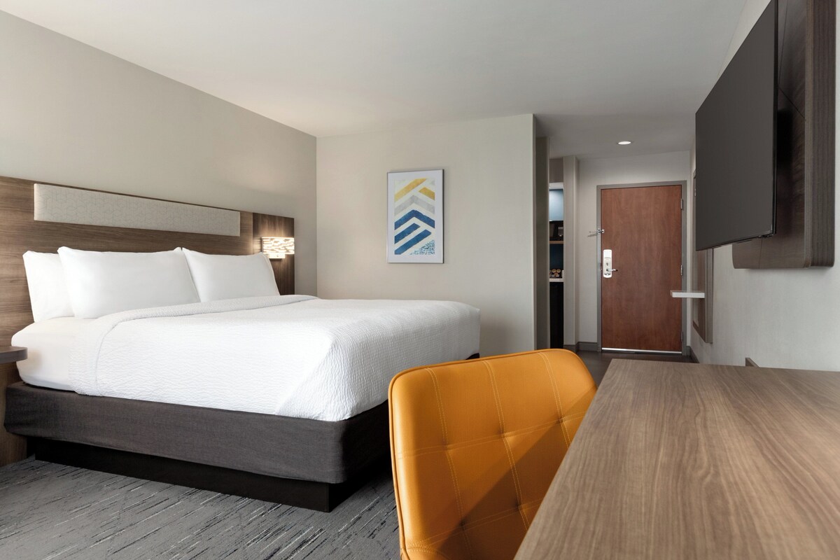 A hotel room with a bed, a desk, a chair, a TV, and a door.