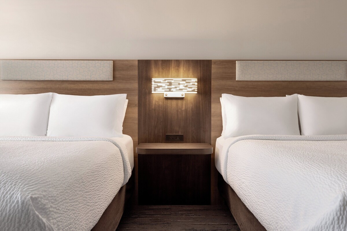 A hotel room with two beds, a nightstand, and a wall-mounted light fixture.