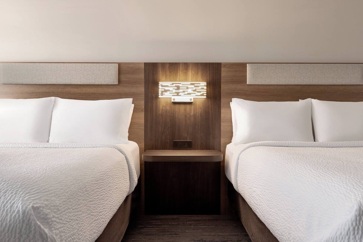 A hotel room with two beds, a nightstand, and a wall-mounted light fixture.