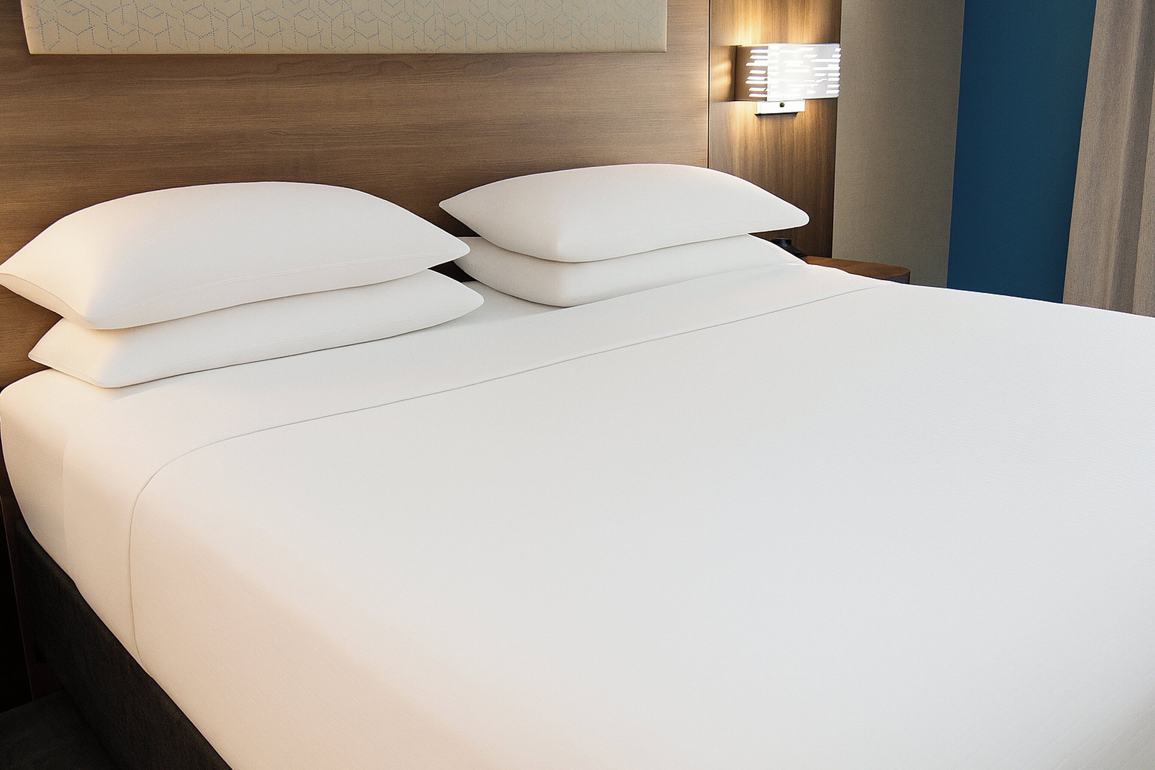 A neatly made bed with white linens and pillows, a wooden headboard, and a wall-mounted lamp.