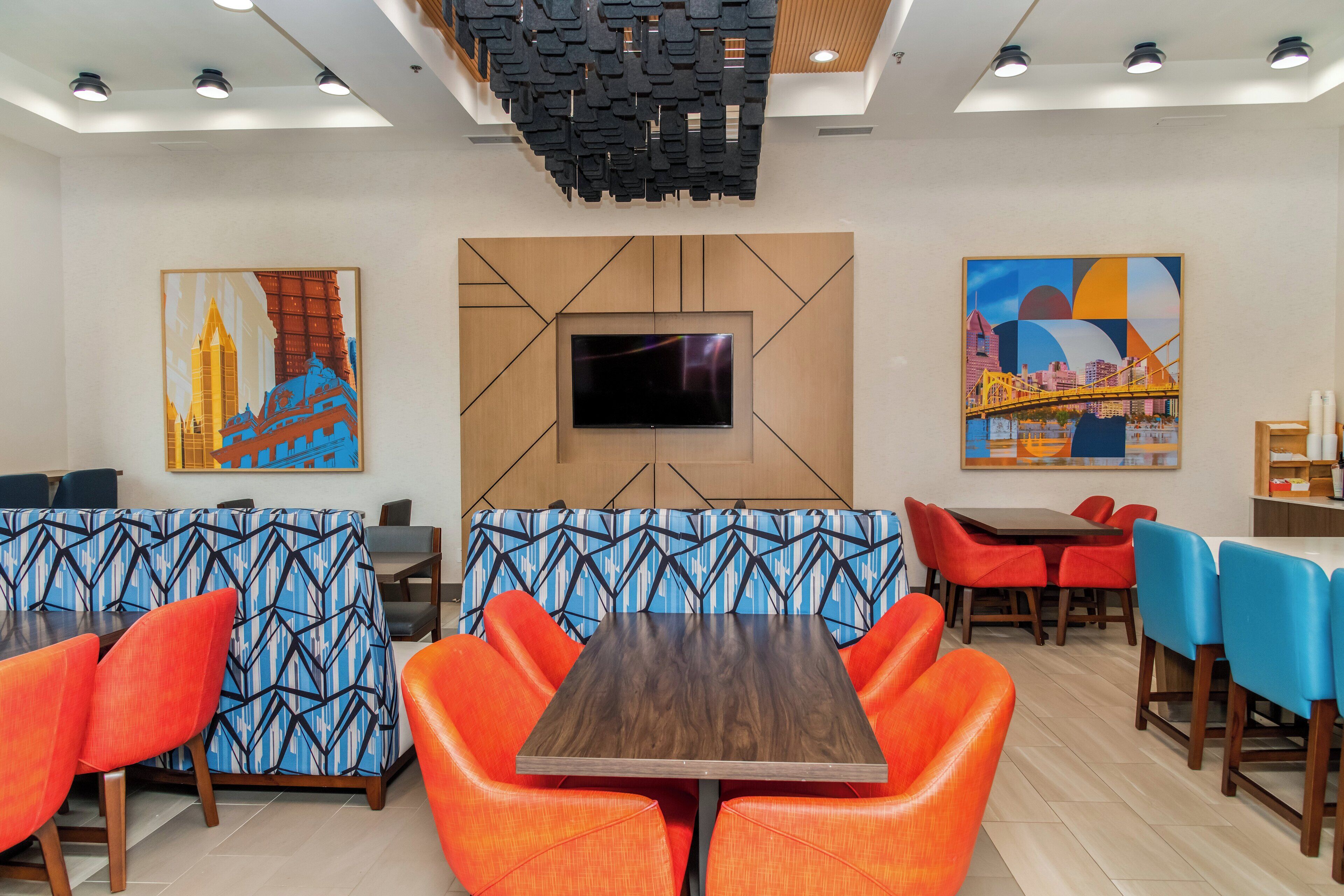 A modern restaurant with a central wooden table, orange chairs, and blue patterned booths.