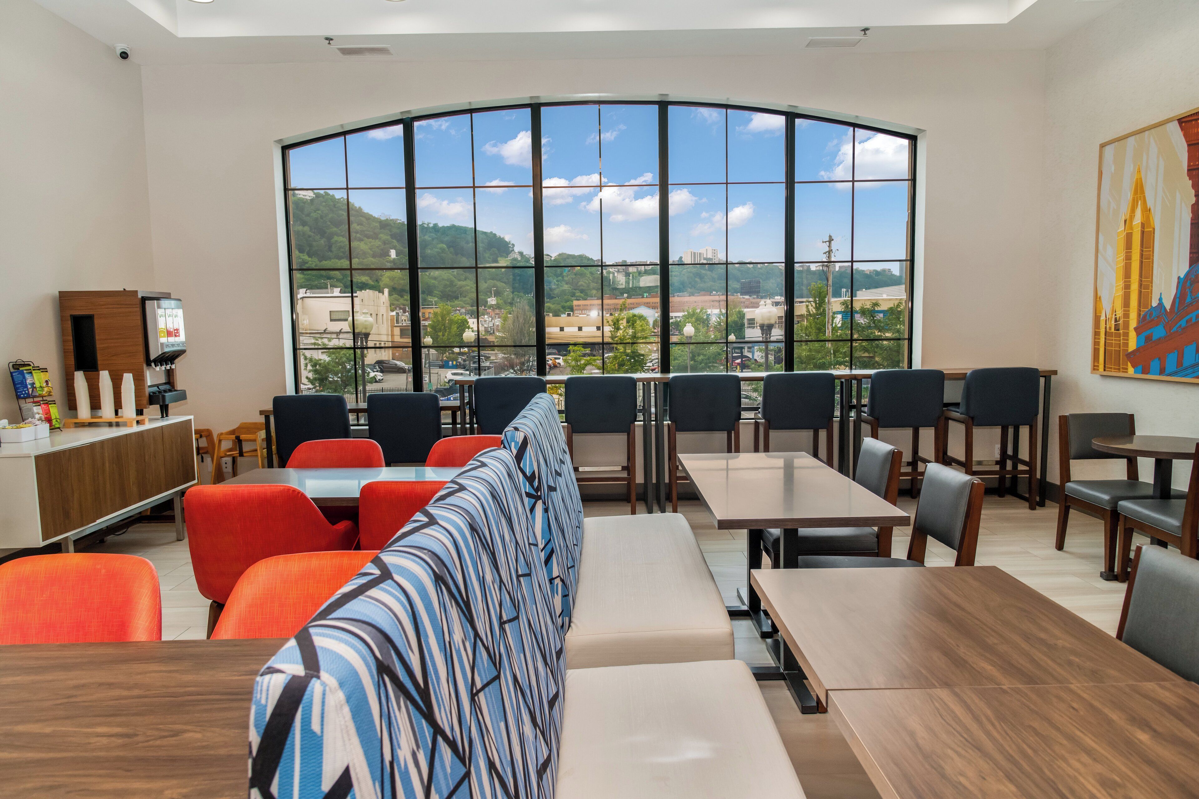 A modern restaurant with a large window offering a view of a cityscape and hills. There are multiple seating areas with tables and chairs, and a counter with a menu display.
