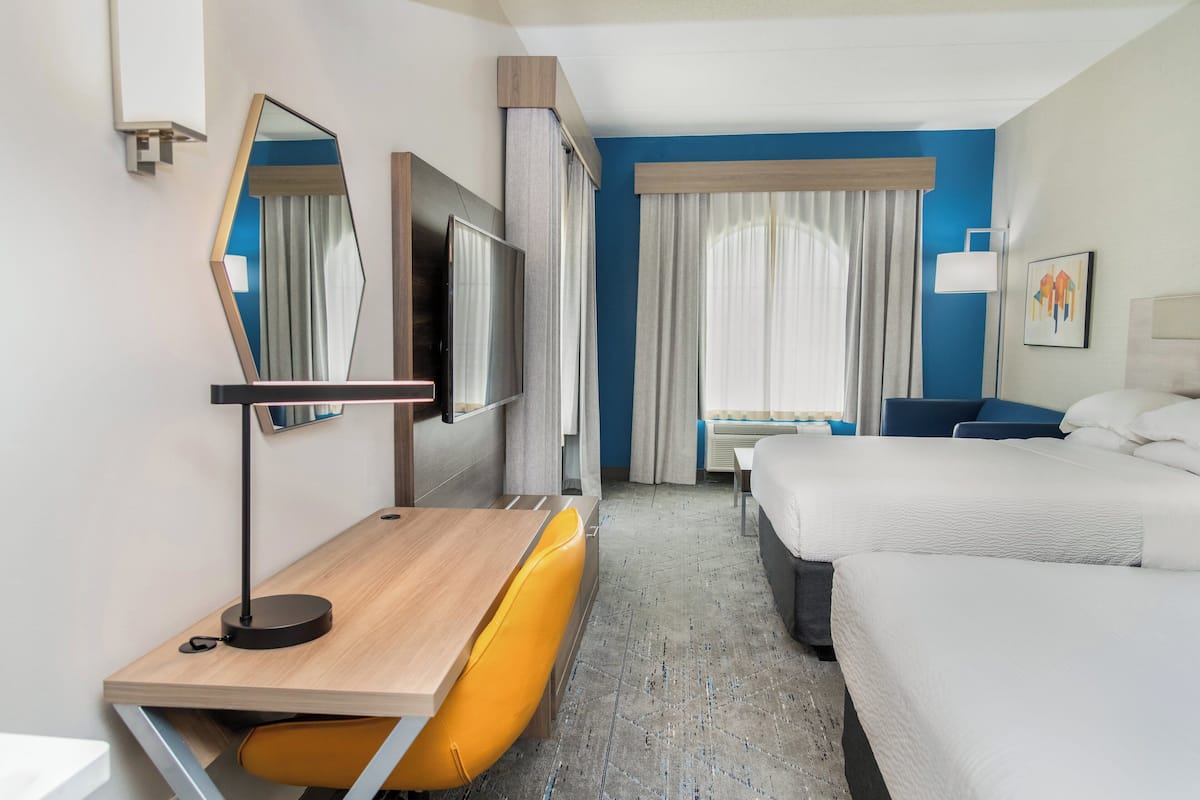 A hotel room with a wooden desk, a yellow chair, a bed, a TV, and a large window with curtains.