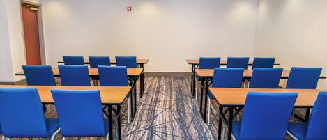 A conference room with rows of blue chairs and wooden tables, a patterned carpet, and a white ceiling with recessed lighting.