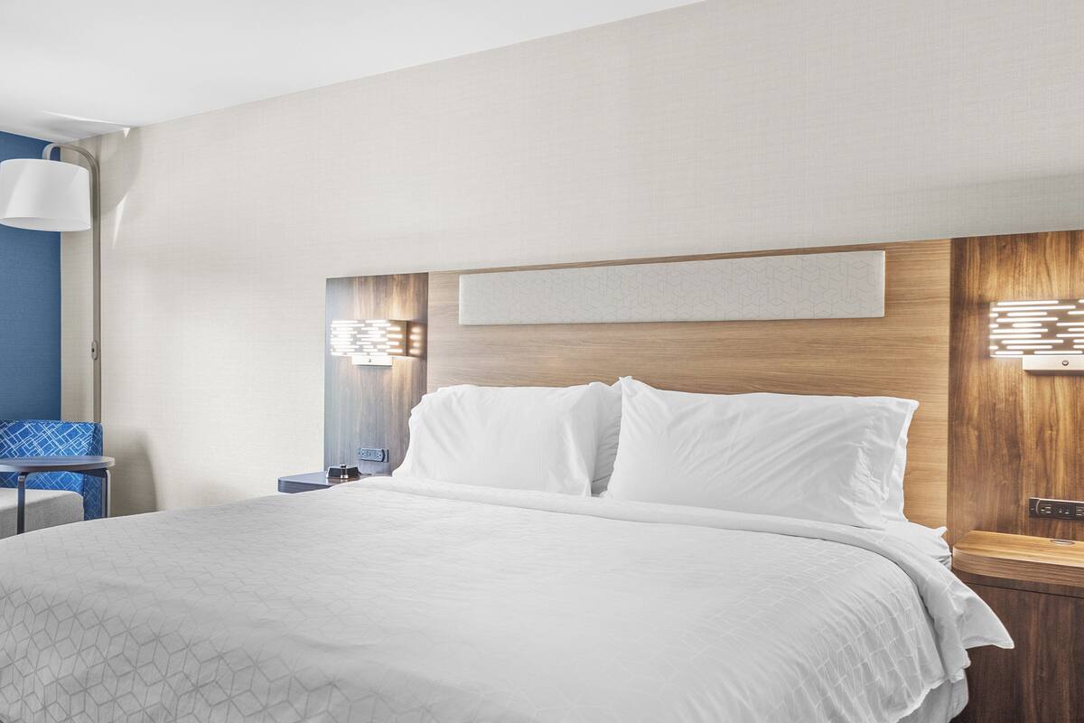 A hotel room with a large bed, a seating area with a blue chair, a lamp, and a wall-mounted light fixture.