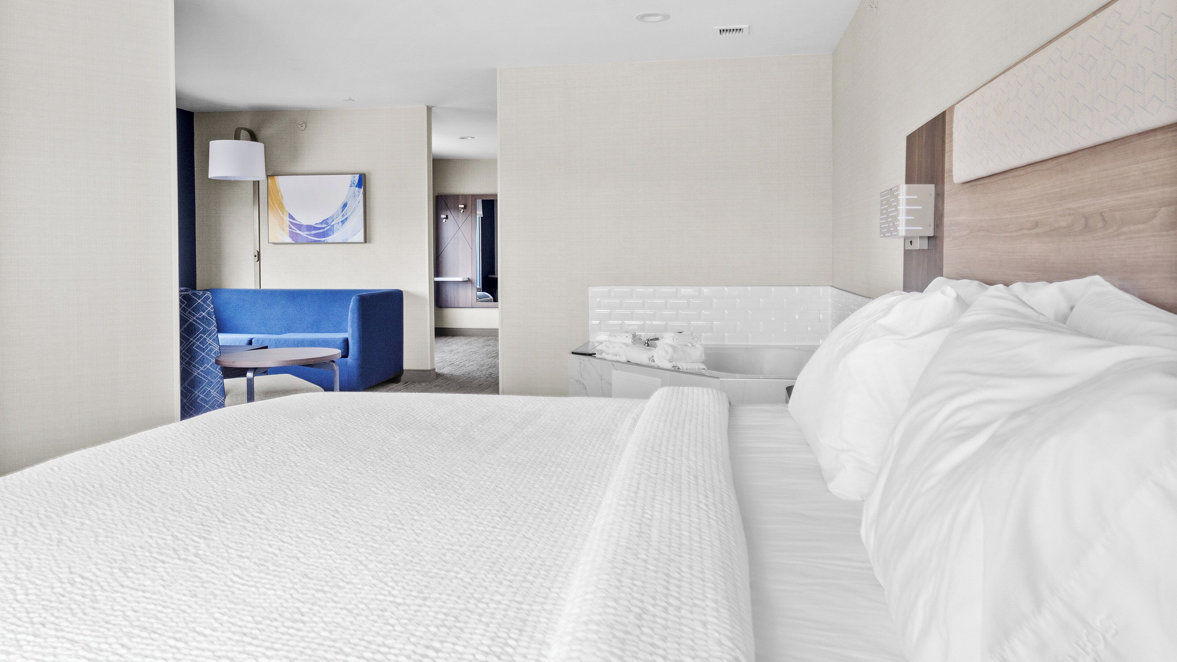 A hotel room with a large bed, a desk with a chair, a bathroom with a mirror and towel rack, and a painting on the wall.