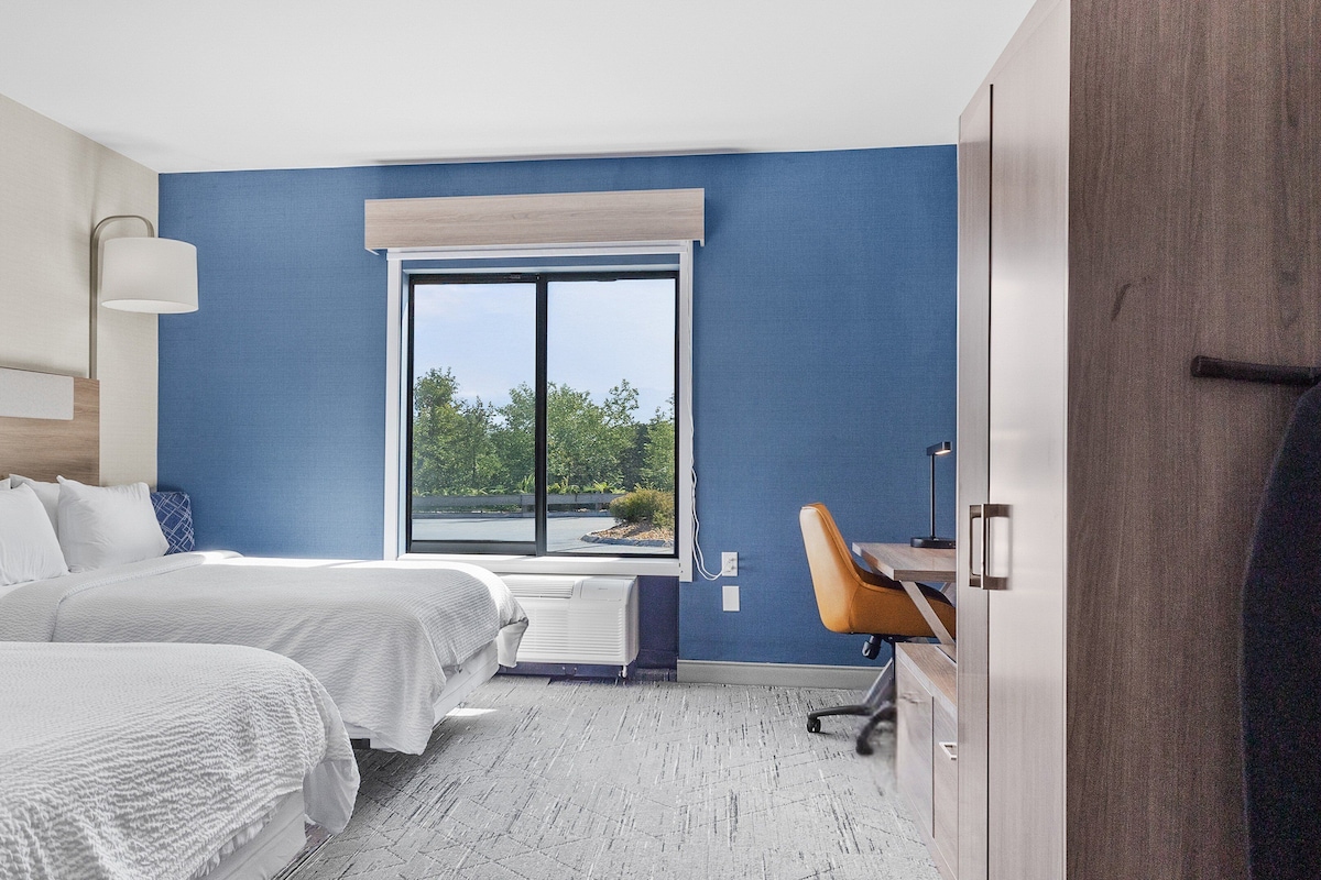 A hotel room with two beds, a large window, a desk, and a chair.