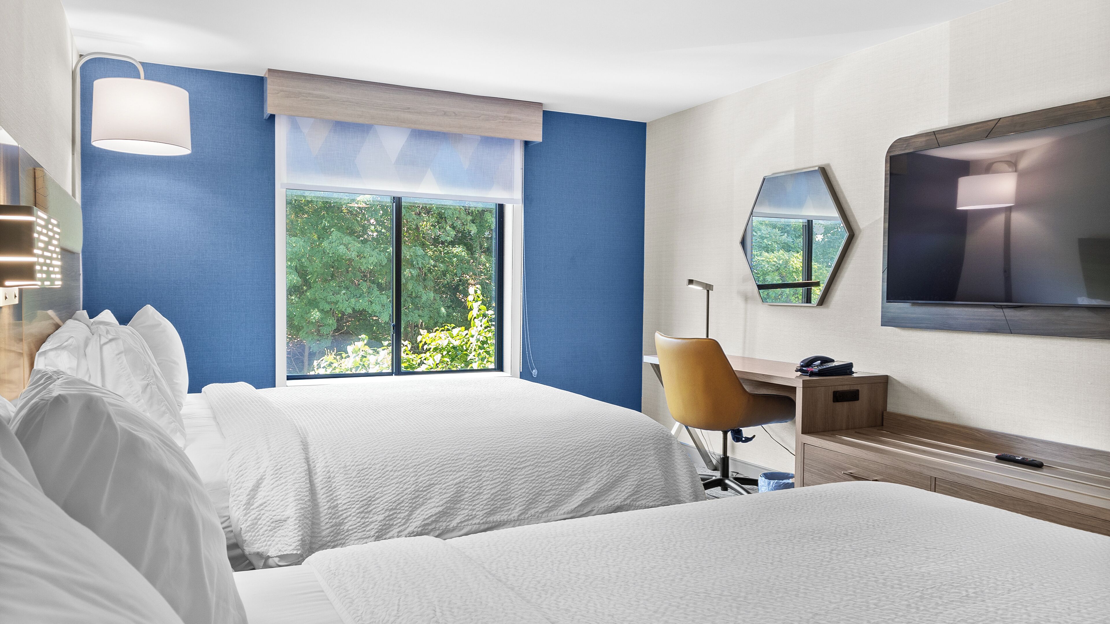 A hotel room with two beds, a desk, a chair, a TV, and a large window with a view of greenery.