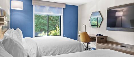 A hotel room with two beds, a desk, a chair, a TV, and a large window with a view of greenery.