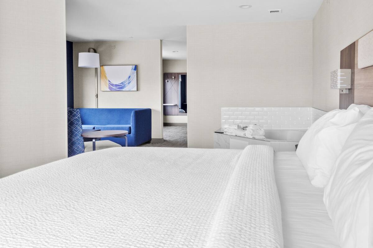 A hotel room with a large bed, a desk with a lamp, a blue chair, and a bathroom with a mirror and towel rack.