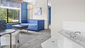 A modern hotel room with a blue sofa, a small table, a window with a view of trees, and a bathroom with a large mirror and sink. - Holiday Inn Express Hotel & Suites Manchester Airport by IHG (Manchester)