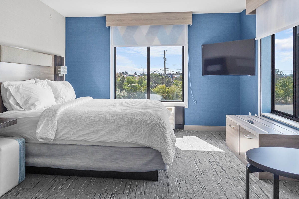 A modern hotel room with a bed, bedside table, TV, and a view of the city.